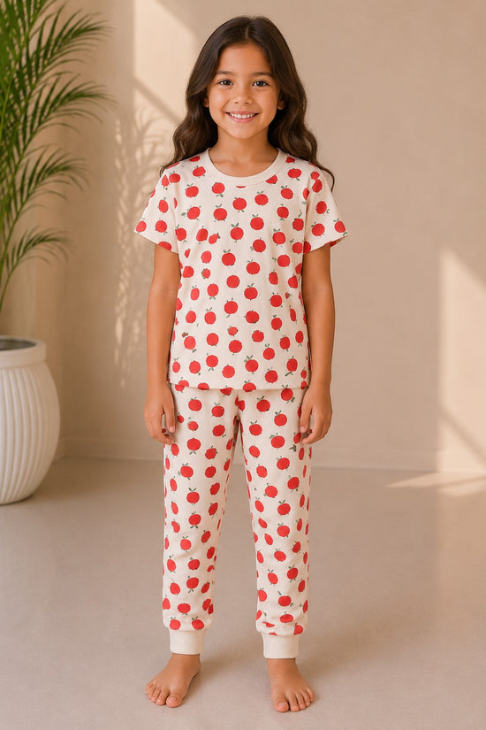 A young girl wearing a Chillpjs cream cotton pajama set with a red apple print, featuring a short-sleeve top and joggers