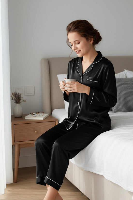 Woman wearing Chillpjs black satin button-down pajama set with white contrast piping, sitting on a bed with a mug.
