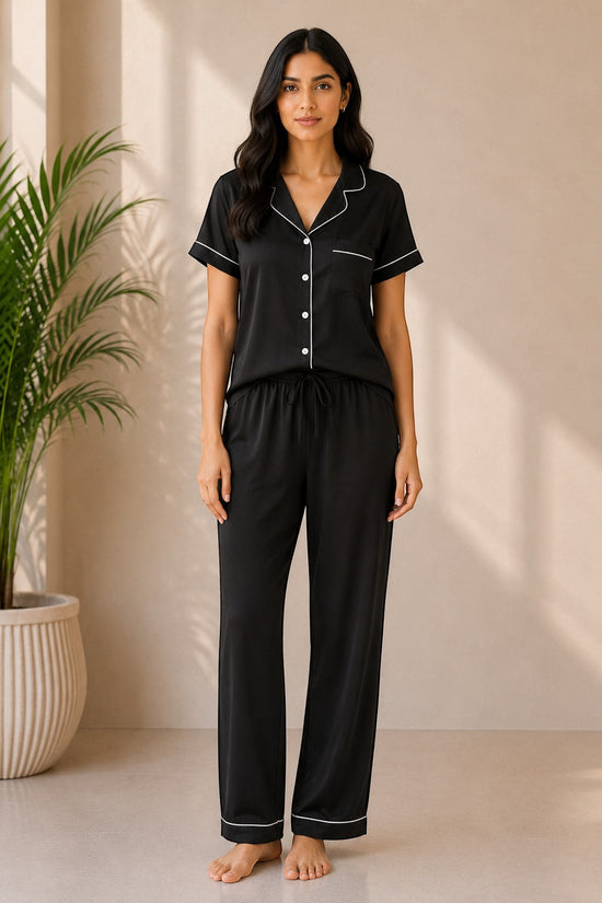 Model wearing a black Chillpjs silk-feel short sleeve pajama set with white piping and drawstring trousers.