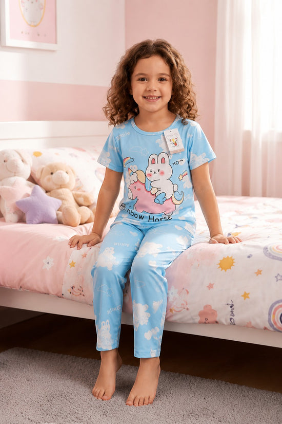 Chillpjs blue cotton night suit featuring a cute rainbow bunny print worn by a smiling girl sitting on a bed.