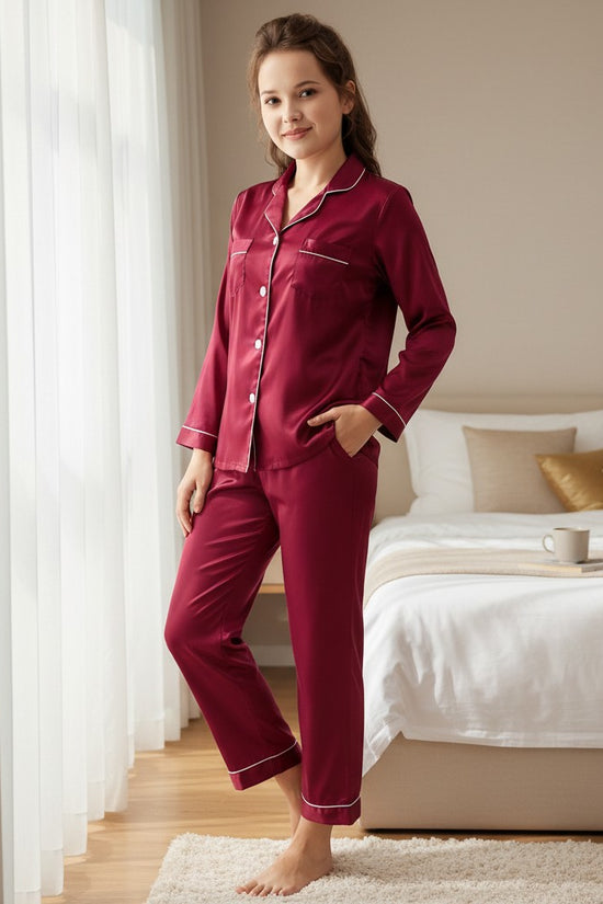 Woman wearing a Chillpjs burgundy satin button-down pajama set with white contrast piping in a bright bedroom