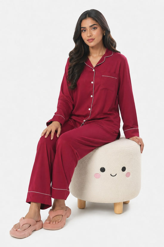 Model wearing a maroon Chillpjs satin silk pajama set with white piping, sitting on a cute cream-colored stool.