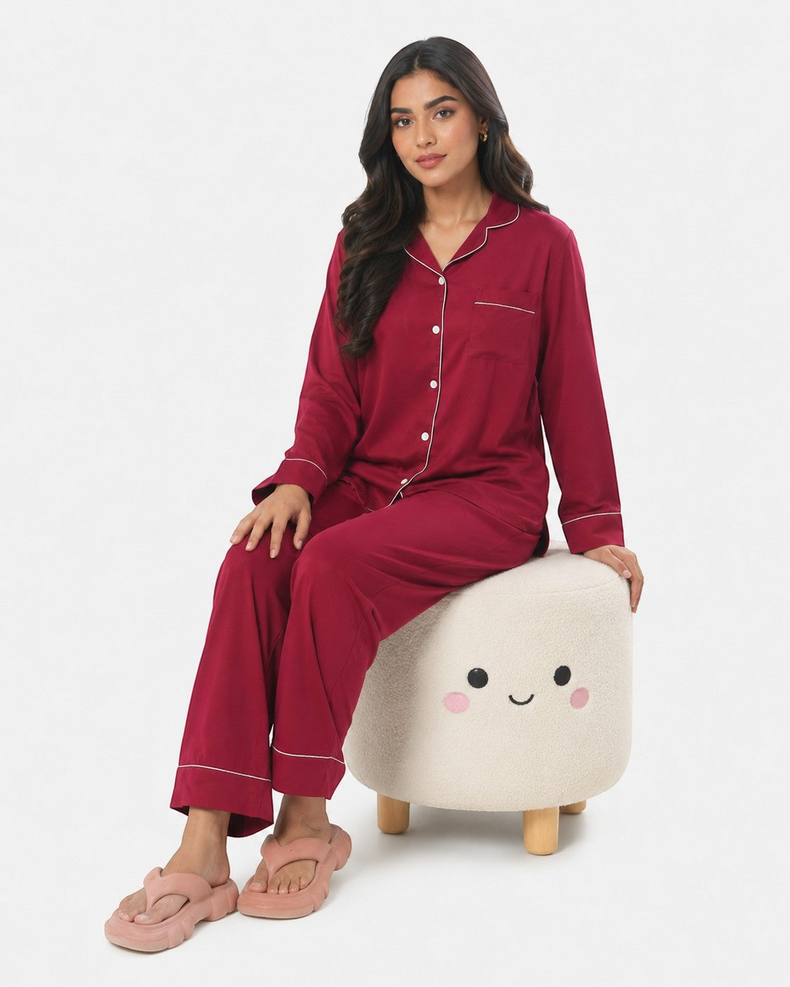 Model wearing a maroon Chillpjs satin silk pajama set with white piping, sitting on a cute cream-colored stool.