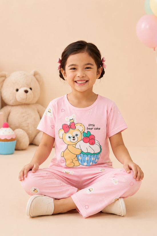 Chillpjs pink cotton night suit with bear and cupcake print worn by a smiling girl sitting on the floor