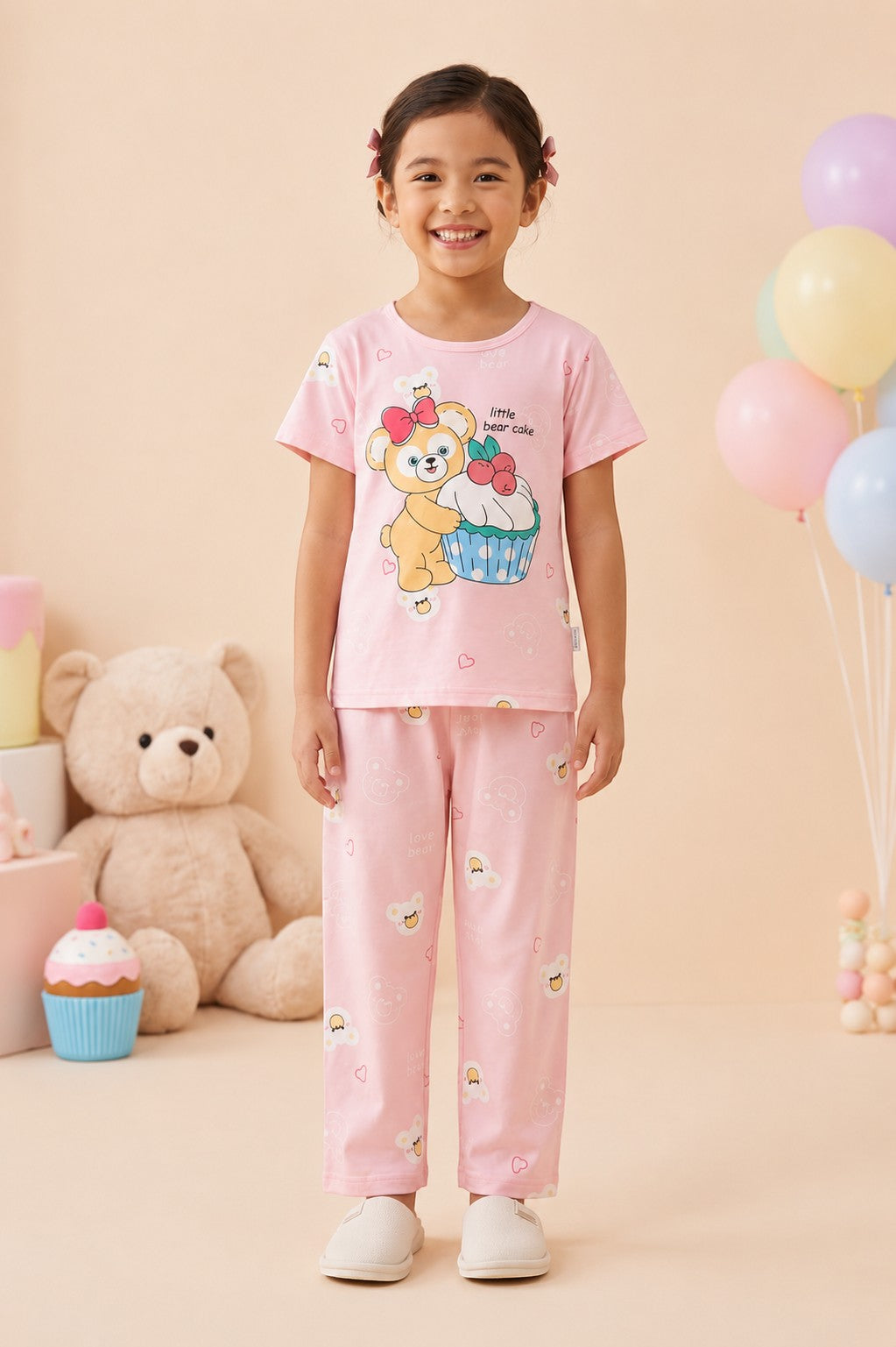 A young girl wearing a Chillpjs pink cotton night suit with a cute bear and cupcake print in a decorated room.